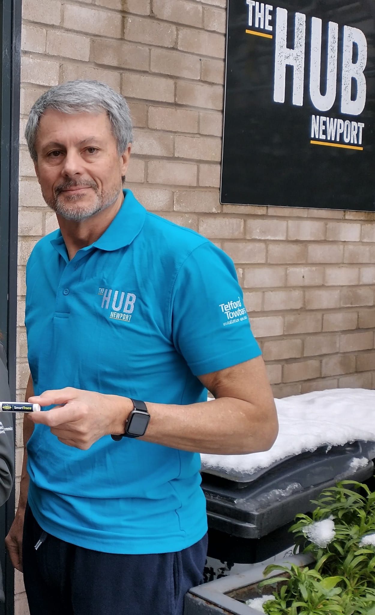 Cllr Tin Nelson wearing a light blue t-shirt with a light coloured brick wall and two snow-covered bins behind him