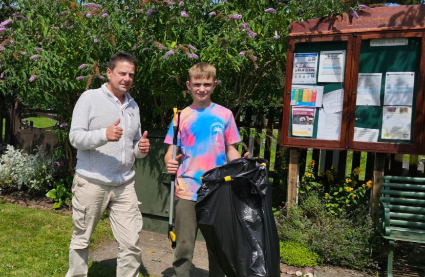 Cllr Steve Burrell with young resident Jake in a garden area. Jake is holding a litter picker and bin bag. The village notice board is in the background.