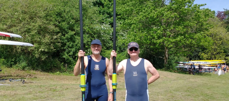 Cllr Tim Nelson with another rower holding an oar each. Both men are standing side by side on grass with trees and a blue sky behind them.