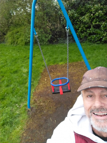 Cllr Tim Nelson selfie in front of refurbished swing. He is wearing a white jacket and beige hat. He is smiling.