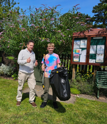 Cllr Steve Burrell with young resident Jake in a garden area. Jake is holding a litter picker and bin bag. The village notice board is in the background.